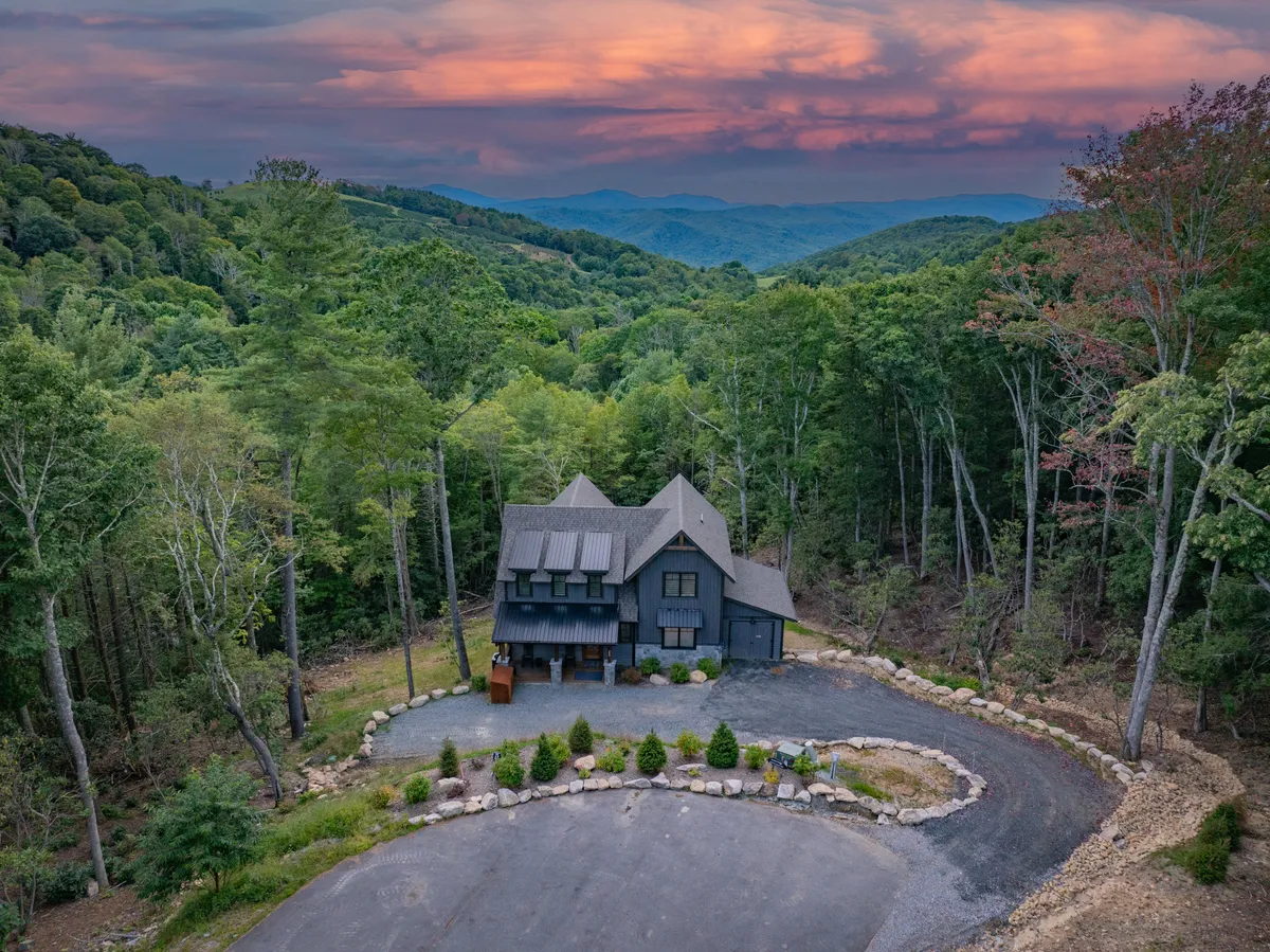 A modern home set among tall trees with mountains on the horizon and a dramatic sunset sky. The expansive landscape invites exploration, with the house blending into the mountainous terrain. The setting suggests easy access to trails and outdoor activities.