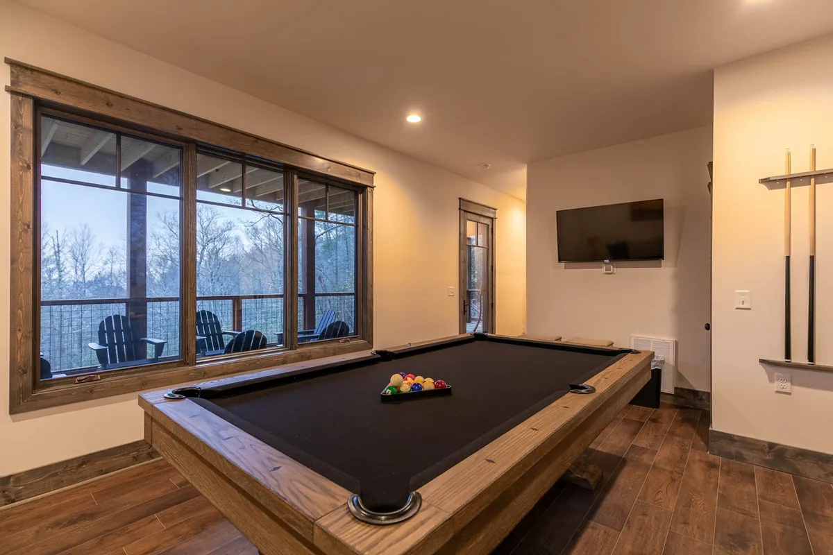 A spacious, well-lit game room featuring a pool table at the center, large windows that let in plenty of natural light, a television mounted on the wall, and modern wooden accents. The seating area nearby suggests a casual, inviting place for both games and relaxation.