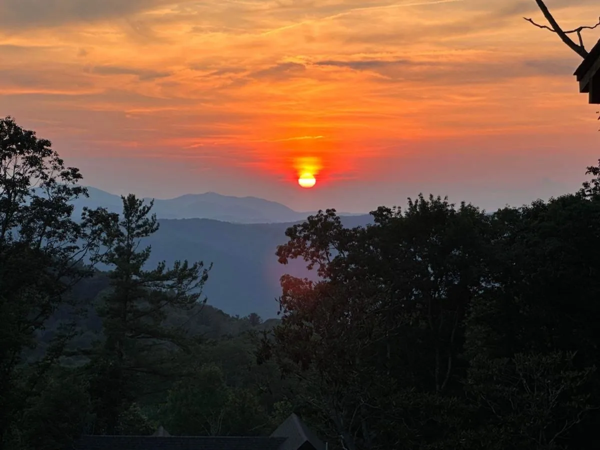 A breathtaking view of a mountain range at sunset, silhouetted trees framing the sky awash in warm, tranquil colors. The landscape is peaceful, perfect for reflection and moments of inspiration.
