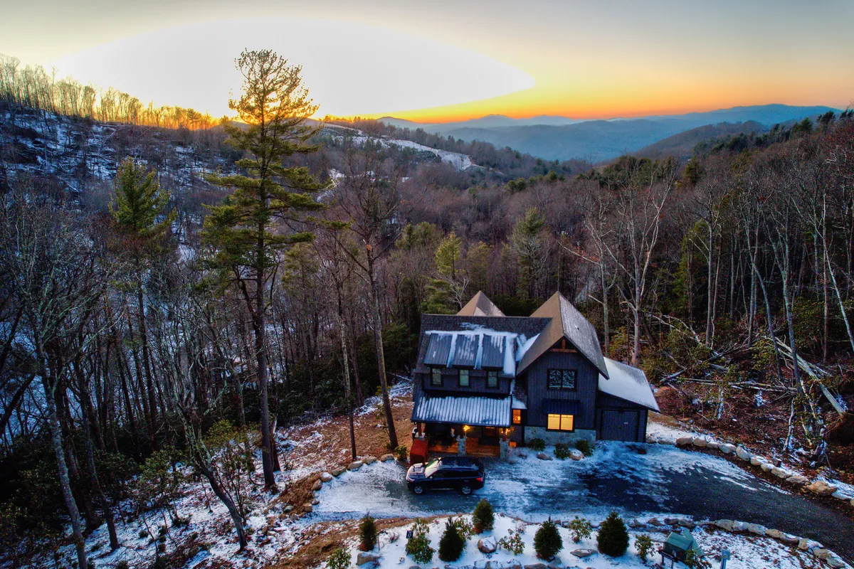 A wide exterior shot showing the house surrounded by trees with a mountain backdrop and a driveway, evoking access to nearby hiking trails and nature walks. A car parked in the driveway hints at the start of mountain adventures, making it easy to envision bringing a dog along for hikes or exploration.