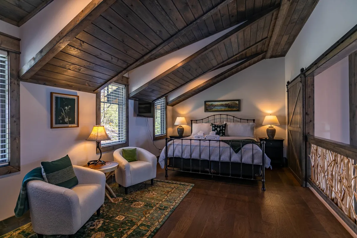 A comfortable bedroom scene with a bed, two accent chairs, lamps on bedside tables, and soft artwork, all arranged in a restful, inviting layout. The mood is tranquil and welcoming.