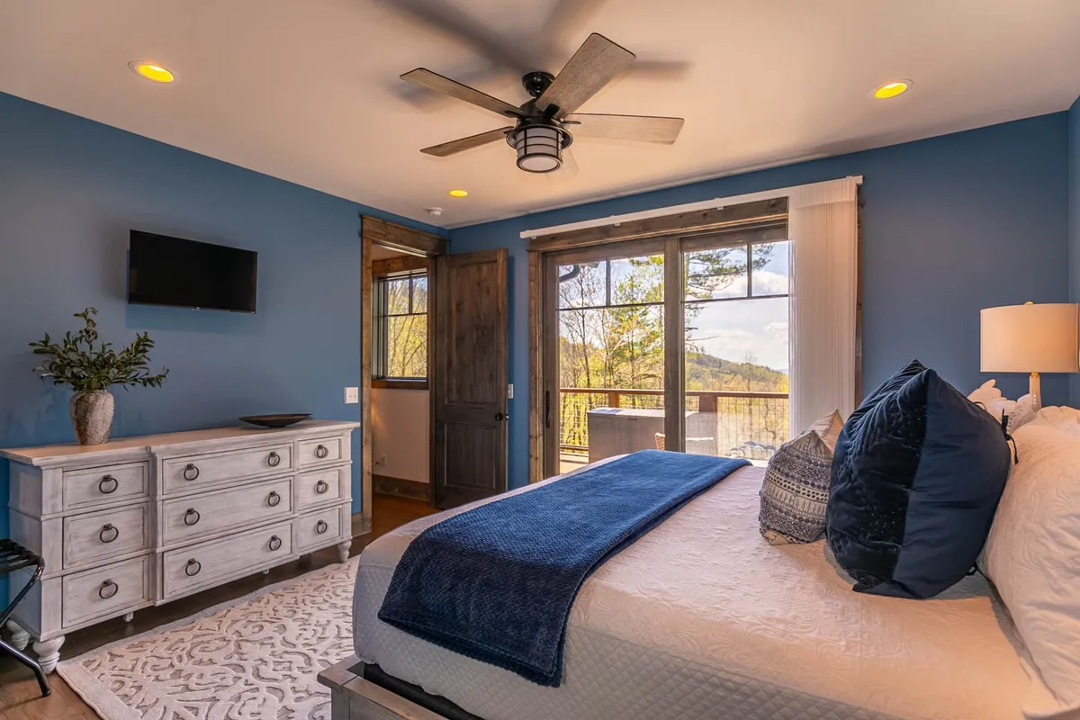 A cozy bedroom with a bed draped in a blue throw, large windows with scenic views, decorative pillows, and a dresser. The room feels restful and offers a touch of mountain character.
