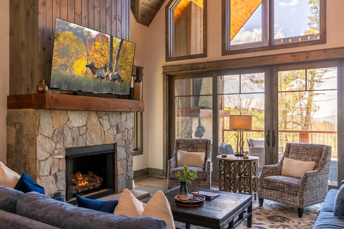 A welcoming living area centered around a fireplace and a large television, framed by expansive windows that bathe the space in natural light. The seating arrangement invites groups to relax for movie nights or fireside chats while enjoying views of the outdoors.