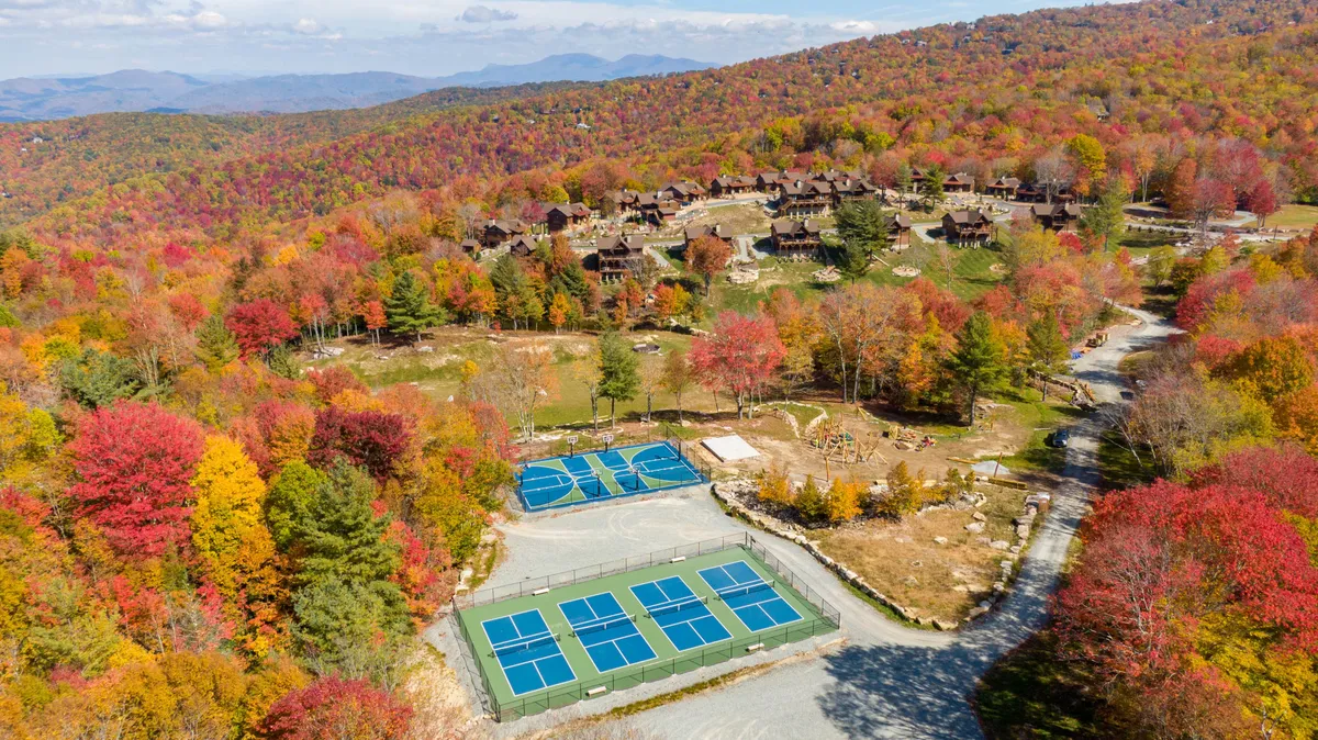 A vibrant exterior scene featuring colorful autumn foliage, tennis courts, scenic cabins nestled among the trees, and a majestic mountain backdrop. The courts are clearly visible and surrounded by natural beauty, suggesting easily accessible sports and recreation amidst the outdoors.