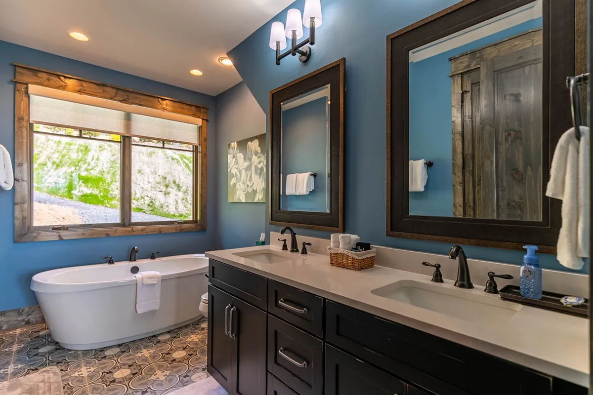 A tranquil bathroom includes a freestanding soaking tub, double vanity with mirrors, large window bringing in natural light, and tasteful wall art. The atmosphere is spa-like and calming.