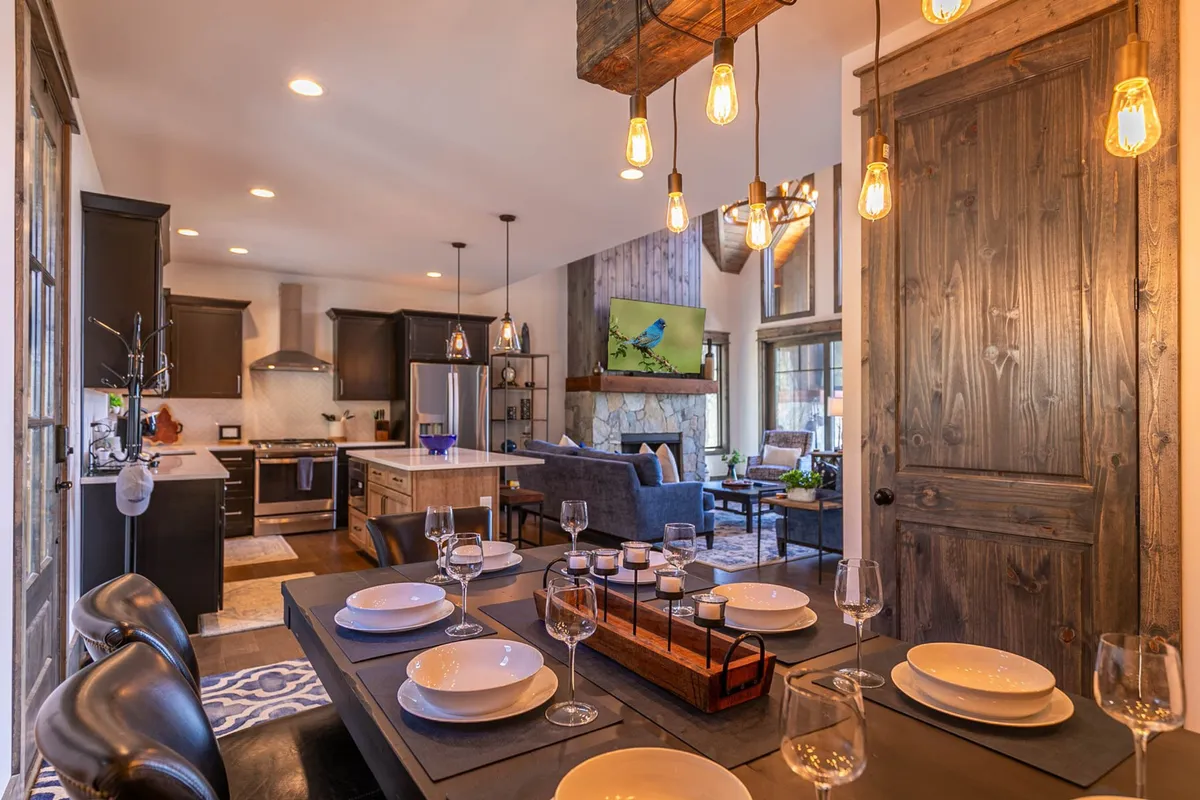 This image displays a seamless open-concept living space where the dining table is set with plates and glasses, the kitchen area is visible in the background, and the living room area includes a cozy seating area around a fireplace with a TV above it. The arrangement creates an inviting environment for gatherings, dining, and relaxing.